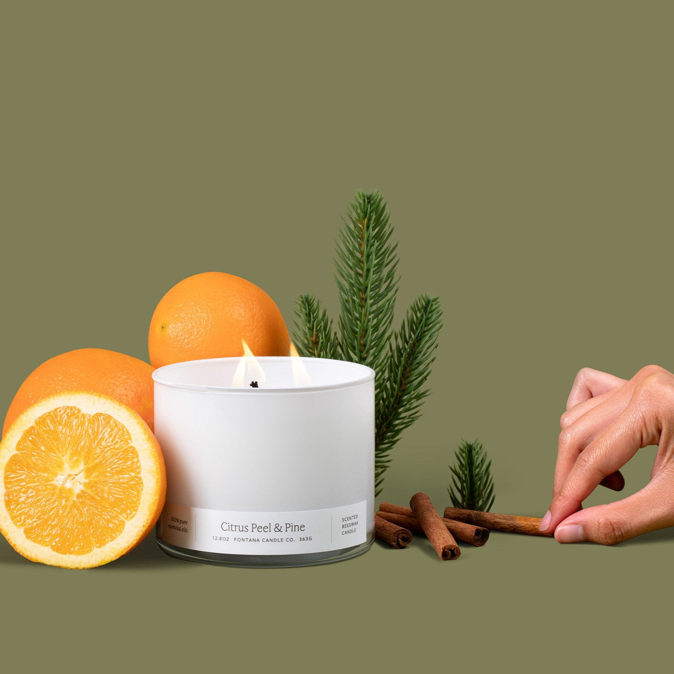 Lit 3-wick candle in a white glass vessel surrounded by oranges, evergreen branches, and cinnamon sticks.