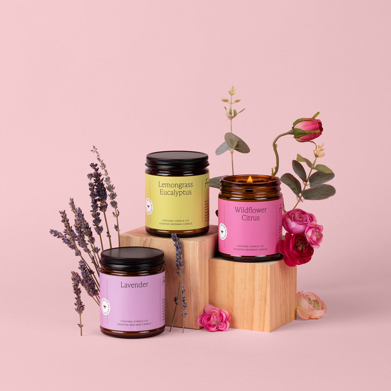 Three amber glass jar candles with bright colorful labels, on a soft pink background, surrounded by Lavender and roses.