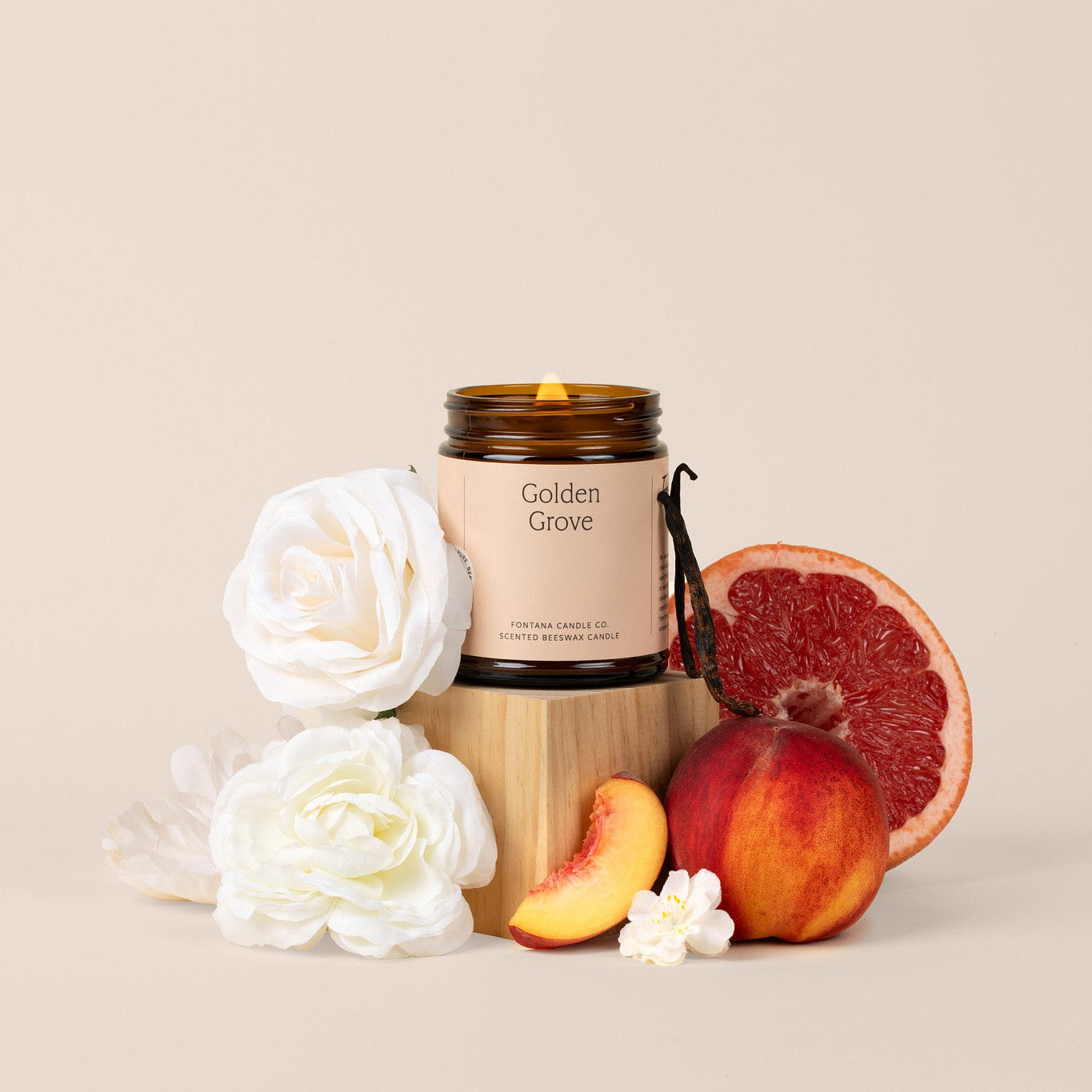 Lit amber glass jar candle labeled 'Golden Grove' with flowers and peaches and grapefruit on a beige background