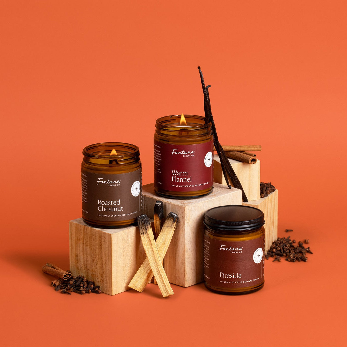 Three amber glass candle jars with labels, styled on wooden blocks against a solid orange background. The candles are labeled 'Roasted Chestnut', 'Warm Flannel', and 'Fireside', with vanilla beans, cinnamon sticks, and cloves used as props.