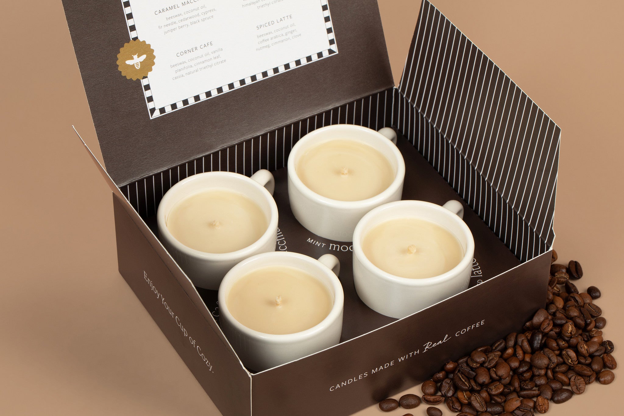 Four white candles in the shape of coffee cups inside a brown box with coffee beans on a beige background.