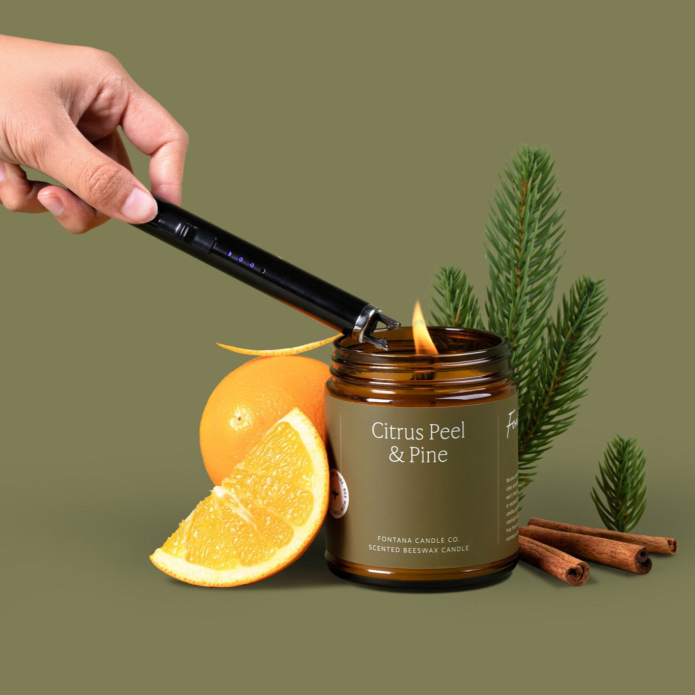 Lit amber glass jar candle with a khaki green label, surrounded by orange peels, fir needles, and cinnamon sticks.