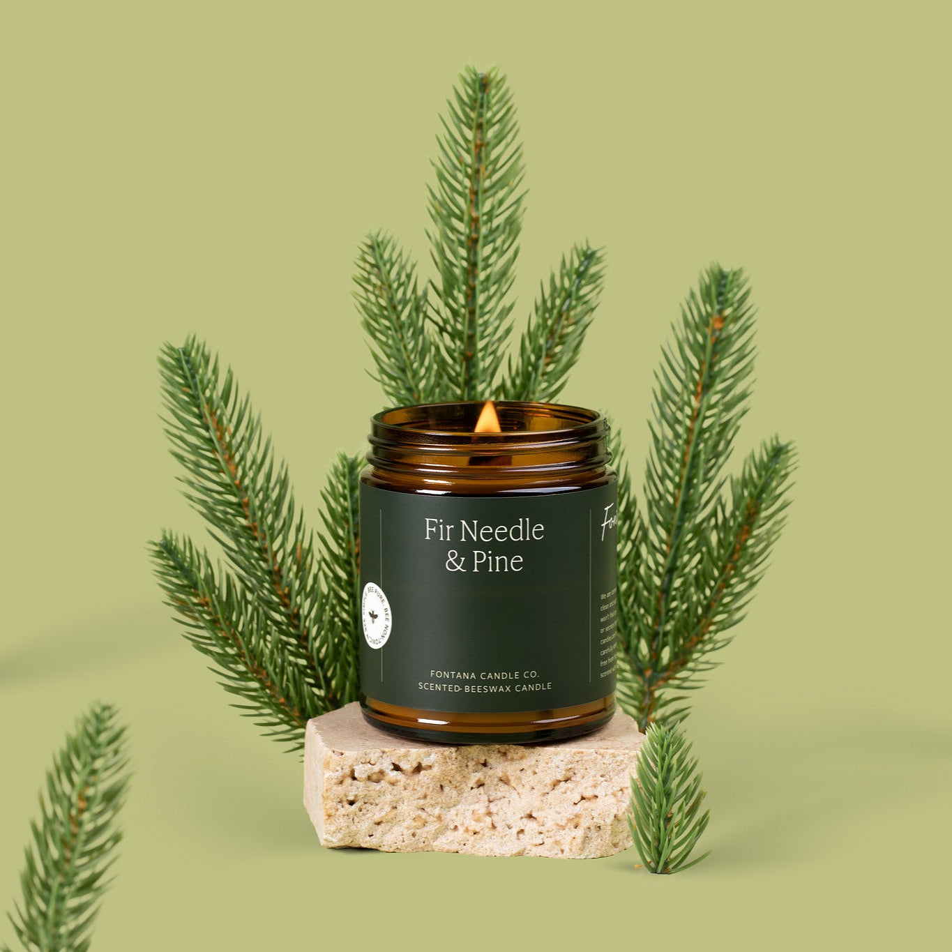 Lit jar Candle labeled ' Fir Needle & Pine' surrounded by pine branches on a light green background.