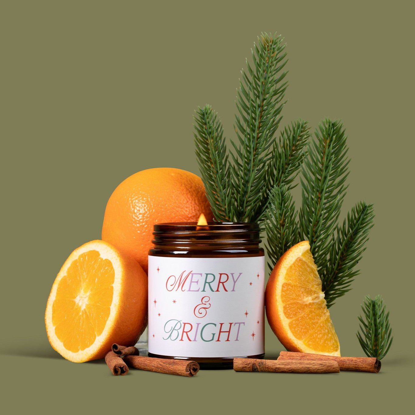 Lit amber glass jar candle labeled 'Merry & Bright' in a festive text with oranges, cinnamon sticks, and pine branches on a green background.