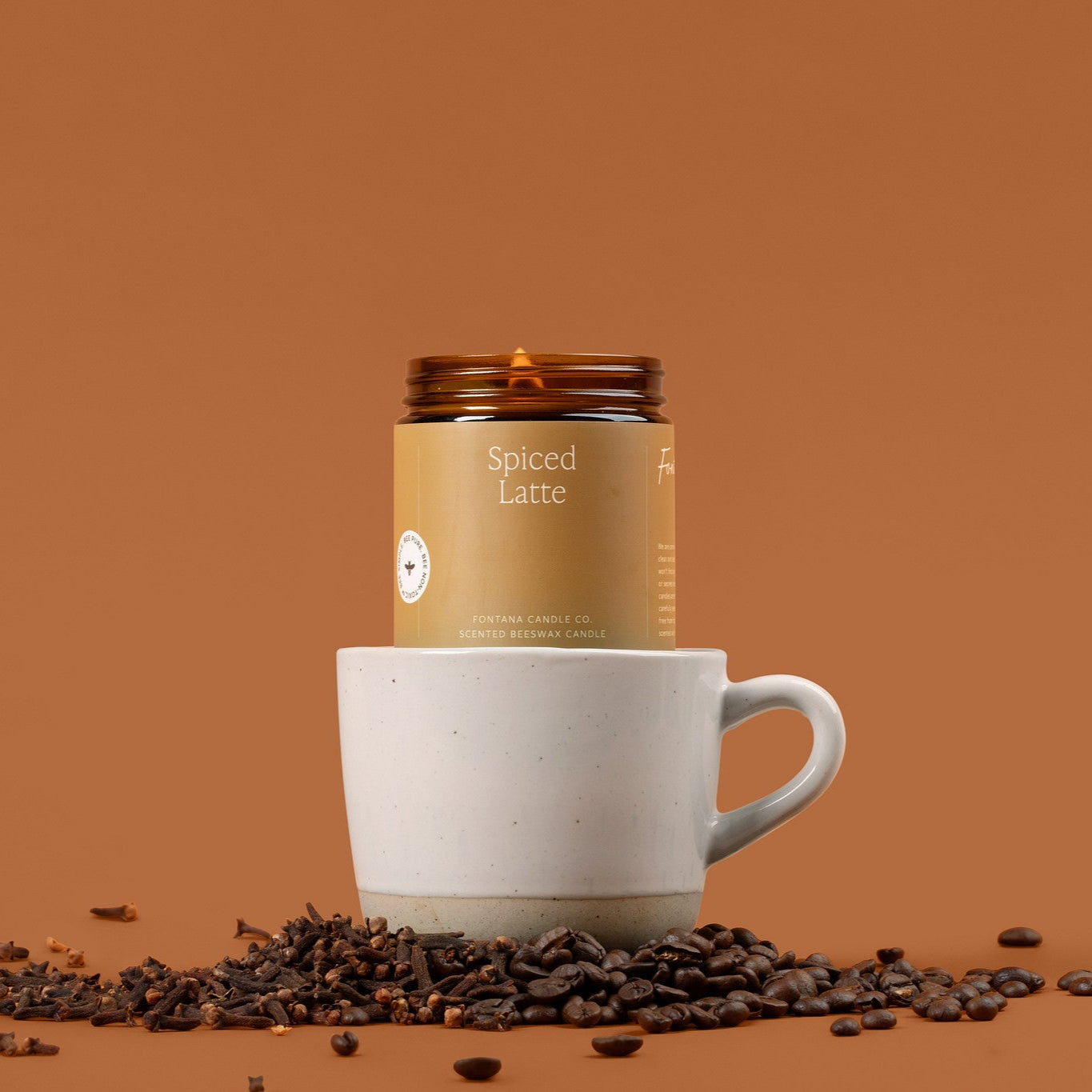 Spiced Latte coffee amber glass jar candle with a white mug and scattered coffee beans on a brown background.