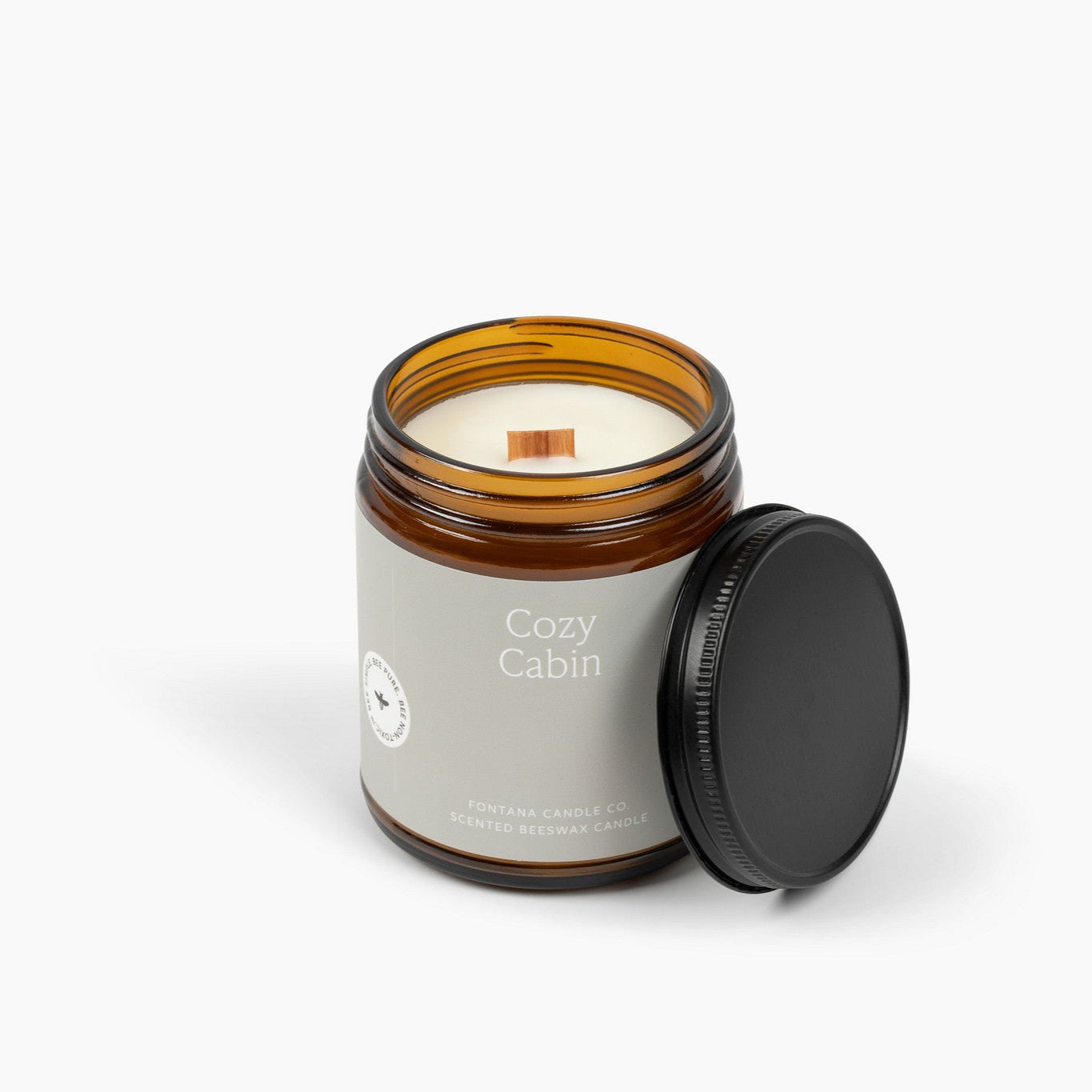 Unlit amber glass jar candle with a light gray label showing the wooden wick and the scent Cozy Cabin.