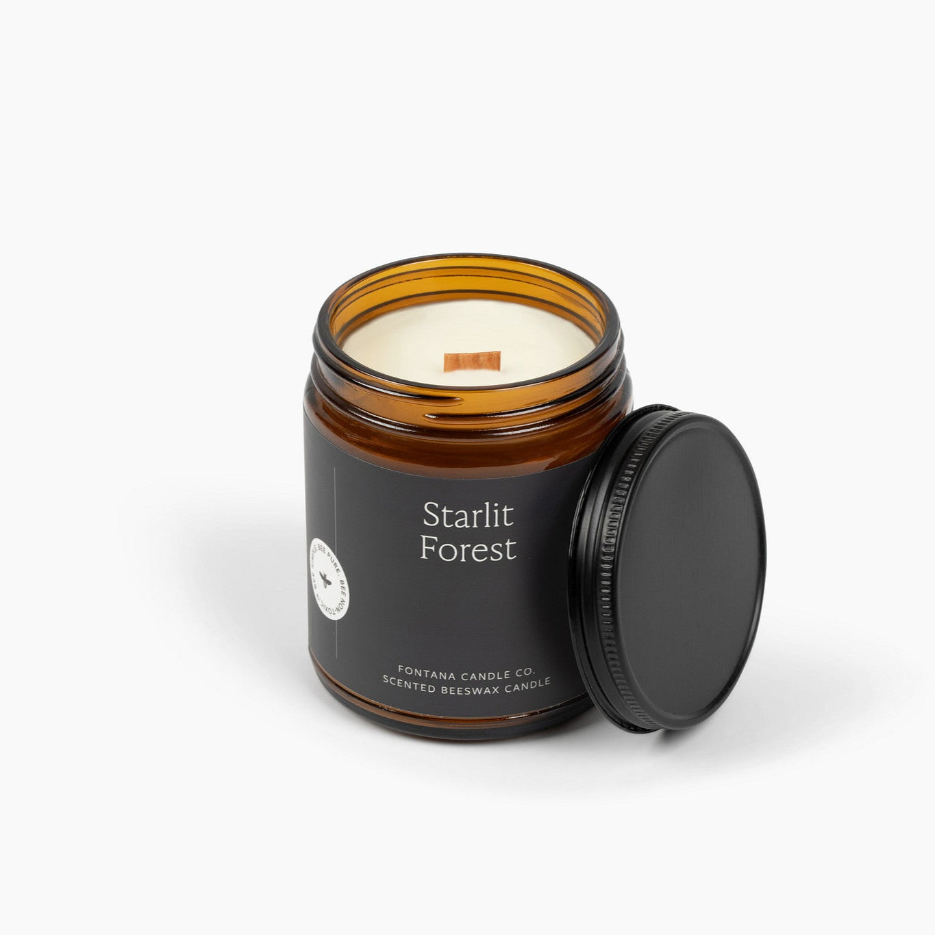 Unlit amber glass jar candle with a dark green label for the scent Starlit Forest and a wooden wick.