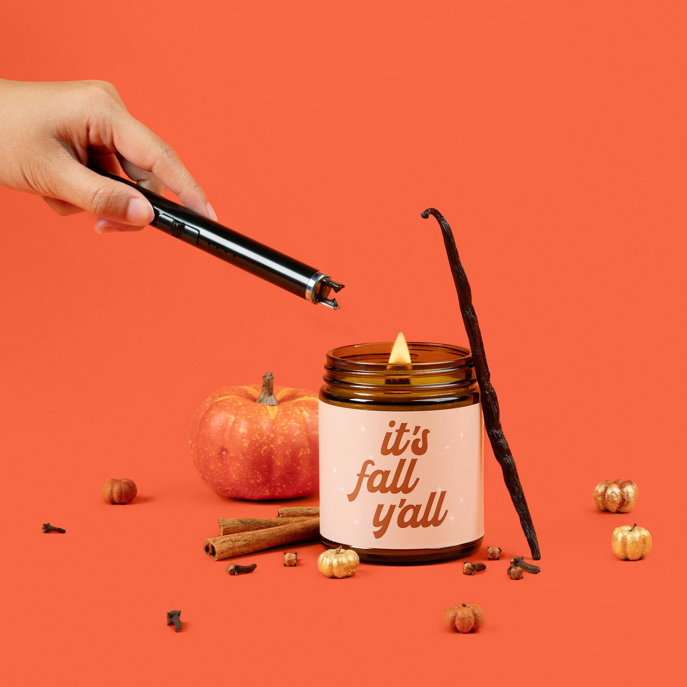 Amber glass jar candle labeled 'it's fall y'all' being lit with autumn-themed elements on an orange background