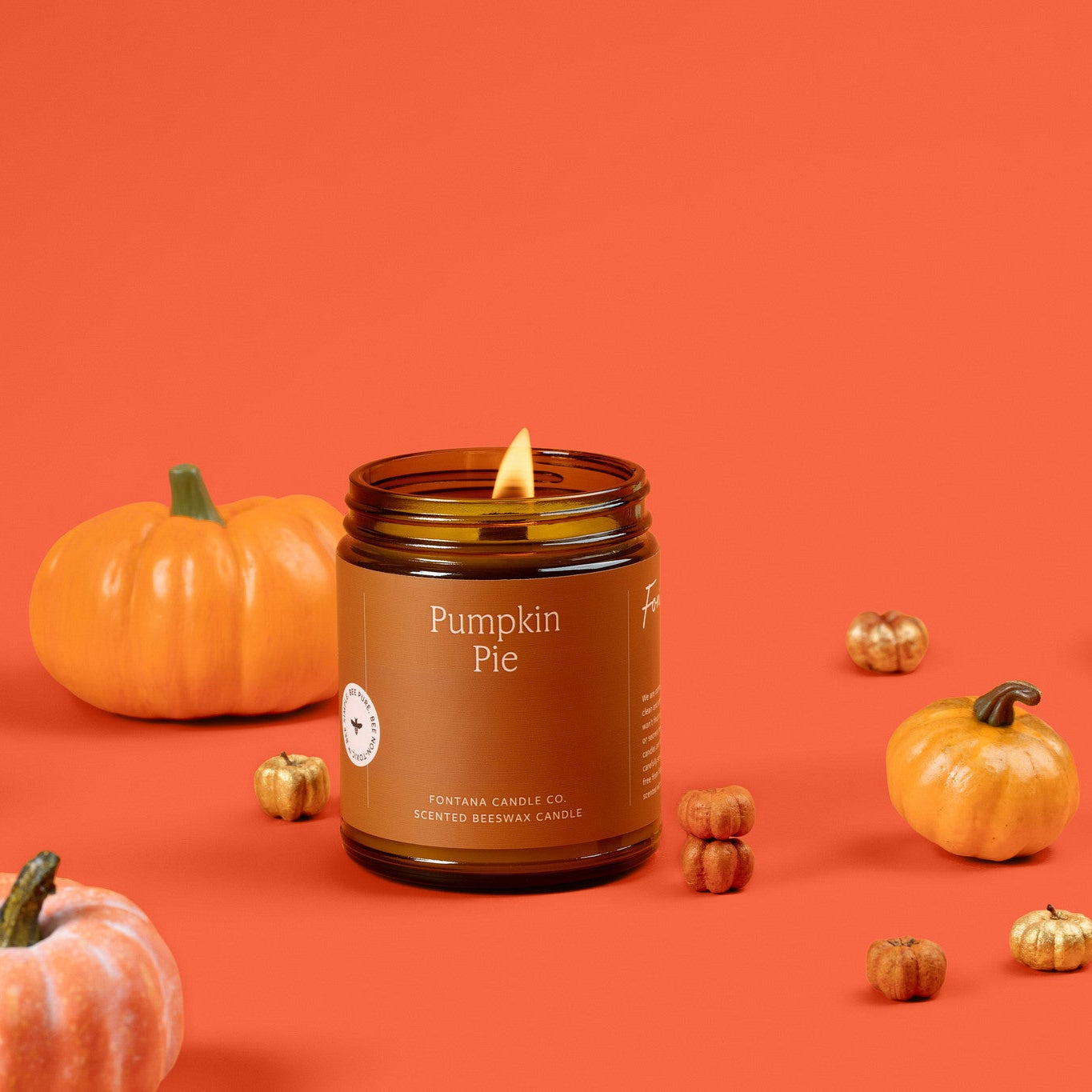 Lit amber glass Pumpkin Pie jar candle with pumpkins on an orange background.